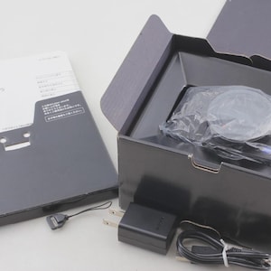 May include: A black Sony digital camera inside a black cardboard box, accompanied by a black power adapter, a black USB cable, a small black wrist strap, and a white instruction manual with the Sony logo and Japanese text printed on the cover.