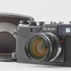 Może przedstawiać: Czarny aparat dalmierzowy Nikon ze srebrnym pierścieniem obiektywu i brązowym skórzanym etui. Obiektyw posiada napis Nippon Kogaku Japan No. 207095. Mały czarny okrągły dekielek obiektywu znajduje się przed korpusem aparatu.