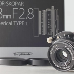 May include: A black Voigtländer Color-Skopar 28mm F2.8 Aspherical Type I camera lens with white text markings, aperture rings, and a focus tab. The lens is positioned in front of its original black and grey product box with matching text.