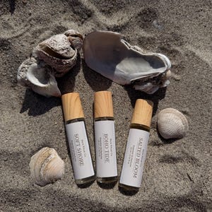 May include: Three essential oil perfume bottles with wooden caps and white labels, labeled "Soft Shore", "Boho Tide", and "Salted Bloom", are arranged on sand with seashells. Each bottle contains 10 ml / 0.33 fl oz of perfume.