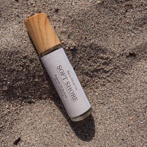 May include: A clear glass essential oil perfume bottle with a light wood cap, labeled "SOFT SHORE" and "Bare Ritual Co." The bottle is 10 ml / 0.33 fl oz. The bottle rests on a sandy surface.