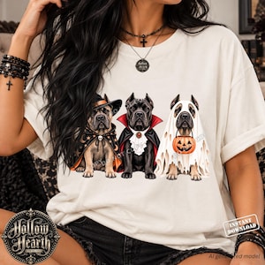 May include: A cream-coloured t-shirt featuring a graphic of three dogs dressed in Halloween costumes. One dog wears a witch hat and cape, the middle dog wears a vampire cape with a ruffled collar, and the third dog is draped in a ghost sheet holding a pumpkin.