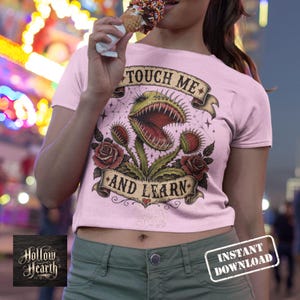 May include: A light pink short-sleeved t-shirt featuring a graphic of a green Venus flytrap with red accents, surrounded by red roses and banners with the text "TOUCH ME" at the top and "AND LEARN" at the bottom, printed in a dark ink style.