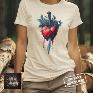 May include: A cream-colored t-shirt featuring a graphic illustration of a red heart pierced by a dark sword, surrounded by dark blue leaves and dripping red liquid. The design is centered on the chest of the shirt, which has short sleeves.