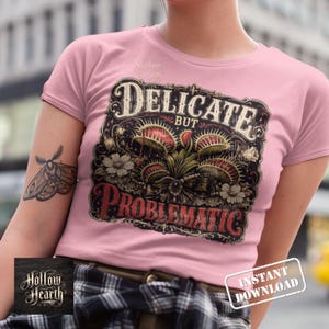 May include: A light pink t-shirt featuring a graphic print of a Venus flytrap plant surrounded by flowers. The text reads "DELICATE BUT PROBLEMATIC" in a decorative font. A moth tattoo is visible on the arm, and a "HOLLOW HEARTH" logo is present.