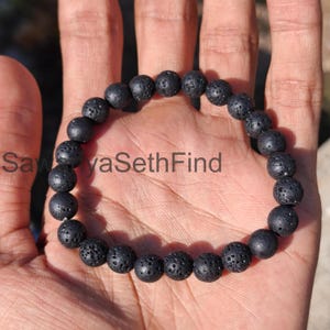 May include: A black lava stone bracelet with round, porous beads. The bracelet is displayed on a hand, with the text "SawyaSethFind" visible. The beads are approximately 1 cm in diameter, strung together to form a complete circle.