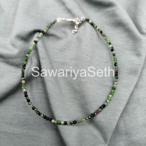 May include: A delicate beaded necklace featuring small, round beads in shades of green, black, and maroon. The necklace is secured with a silver clasp. The text "SawariyaSeth" is visible in white.