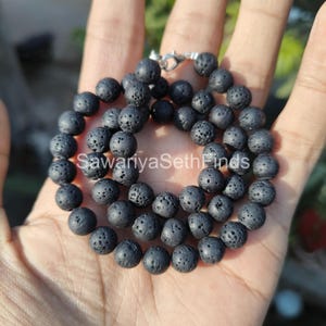 Pode incluir: Um colar de pedra de lava preta com fecho prateado. O colar é composto por pequenas contas redondas e porosas. O texto "SawariyaSethFinds" é visível na imagem. O colar é exibido em uma mão.