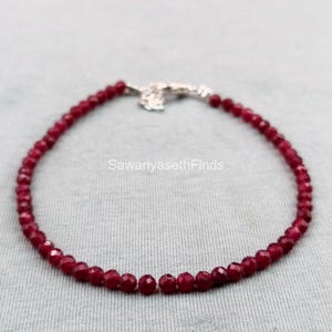 May include: A delicate bracelet featuring small, faceted, deep red beads. The beads are strung together, forming a complete circle, and are secured with a silver-colored clasp. The bracelet is displayed on a light gray surface.
