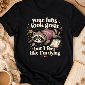 Peut inclure: T-shirt noir avec un dessin d'un raton laveur portant un t-shirt rose avec l'inscription "Chronically Over It". Le texte "your labs look great but I feel like I'm dying" est également imprimé. Le raton laveur est à côté d'un presse-papiers.