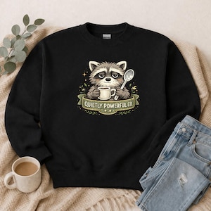 May include: Black sweatshirt featuring a raccoon holding a coffee cup and spoon. The raccoon is surrounded by leaves and stars, with the text "QUIETLY POWERFUL CO." on a banner. A coffee cup and folded jeans are in the background.