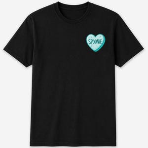 Peut inclure: Un t-shirt noir à manches courtes avec un petit motif en forme de cœur bleu clair sur la poitrine gauche. Le cœur est entouré d'un contour blanc et porte l'inscription SPOONIE en lettres capitales bleu foncé au centre.