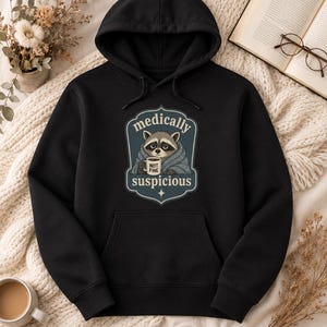 Peut inclure: Sweat à capuche noir avec une poche avant et un motif d'un raton laveur enveloppé dans une couverture, tenant une tasse. Le texte sur le motif indique "medically suspicious" et "not fine".