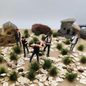 May include: Miniature figures of people in casual attire are scattered across a diorama landscape. The scene includes small stone buildings, green tufts of grass, and scattered rocks, creating a detailed outdoor setting. The figures are engaged in various poses.