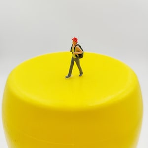 May include: A miniature figure wearing a red hat and backpack walks across a bright yellow, cylindrical object. The figure is dressed in gray pants and a black backpack. The background is a plain white.
