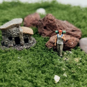 Könnte beinhalten: Miniatur-Szene mit einem Wanderer mit Rucksack, einem kleinen Steinhäuschen und rotbraunen Felsen, eingebettet in grünes Moos. Der Wanderer trägt eine rote Kappe, ein graues Tanktop und eine khakifarbene Hose. Das Haus hat ein Strohdach.