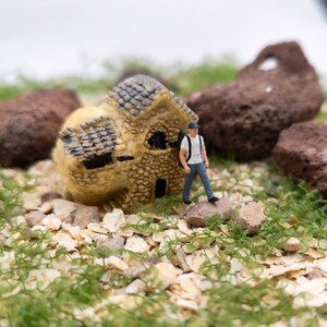 May include: Miniature scene featuring a small stone house with a gray roof and a tiny figure of a person with a backpack. The scene is set on a bed of small rocks and green foliage, with large brown rocks in the background.