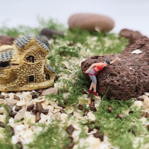 May include: Miniature scene featuring a small stone house, a figure in athletic wear, and a large, textured rock. The scene is set amidst green moss and small stones, creating a whimsical, outdoor setting. The figure appears to be climbing the rock.