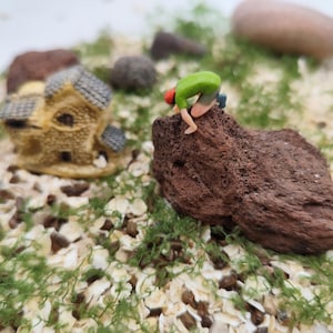 May include: Miniature scene featuring a small house and a figure climbing a brown rock. The figure wears a green shirt, red hat, and tan shorts. The scene is set on a bed of white and tan pebbles with green moss.