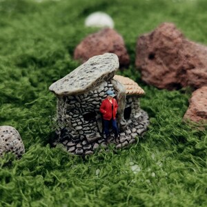 May include: Miniature stone house with a person in a red jacket and blue jeans. The house has a textured roof and is surrounded by green moss and small rocks. A whimsical, detailed diorama.