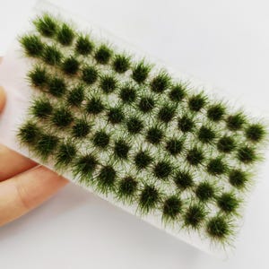 Può includere: Un foglio di ciuffi d'erba verde in miniatura, perfetto per il modellismo o l'artigianato. L'erba è di un verde vibrante, con un aspetto denso e strutturato. I ciuffi sono disposti in file ordinate su un supporto trasparente.