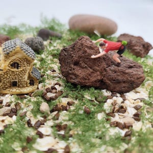 May include: Miniature scene featuring a small house with a stone facade, a person climbing a brown rock, and other rocks. The scene is set on a bed of green moss and small grains, creating a diorama effect.