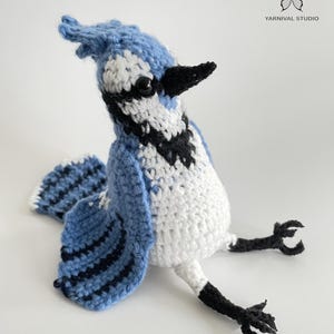 以下が含まれることがあります： 手作りの鉤針編みのアオカケス。白と黒のアクセント。鳥は白い胸、黒い縞模様の青い翼、黒い嘴を持っています。鳥は足を伸ばして座っています。Yarnival Studioのロゴが右上隅にあります。