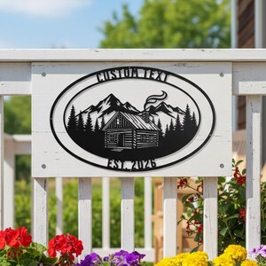 Puede incluir: Letrero de metal negro con un diseño de cabaña, montañas y árboles, dentro de un marco ovalado. El letrero dice "CUSTOM TEXT" encima de la cabaña y "EST. 2026" debajo. Montado en una valla de madera blanca, con flores coloridas en macetas de terracota.