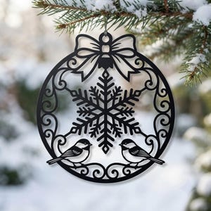 Può includere: Un ornamento circolare nero caratterizzato da un design a fiocco di neve centrale affiancato da due piccoli uccelli nella parte inferiore. L'ornamento è sormontato da un fiocco decorativo e motivi a volute lungo il bordo esterno.