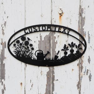 Könnte beinhalten: Schwarzes Metall-Gartenschild mit ovalem Rahmen und dem Text "CUSTOM TEXT". Das Schild zeigt Silhouetten von Sonnenblumen, einem Vogel, einer Gießkanne, einer Schaufel und Wurzelgemüse. Das Schild ist auf einer verwitterten weißen Holzoberfläche montiert.