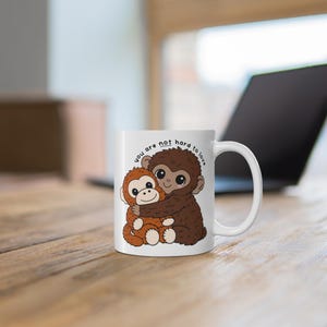 May include: White ceramic mug featuring a cartoon illustration of two hugging monkeys. The monkeys are brown and orange with large eyes. Text on the mug reads "you are not hard to love".