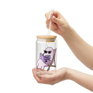 May include: A clear glass tumbler with a bamboo lid and a glass straw. The glass features a graphic of a light purple ghost wearing sunglasses, sitting in a beach chair, and reading a book with the text SPOOKY SUMMER printed on the cover.