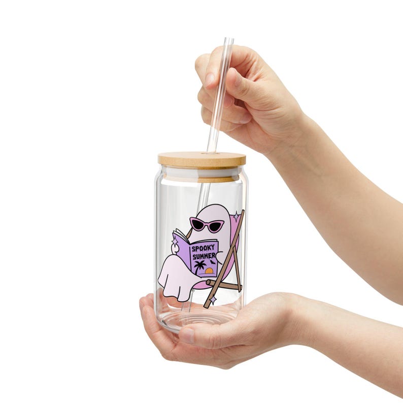 May include: A clear glass tumbler with a bamboo lid and a glass straw. The glass features a graphic of a light purple ghost wearing sunglasses, sitting in a beach chair, and reading a book with the text SPOOKY SUMMER printed on the cover.
