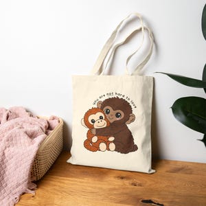 May include: Cream-colored tote bag featuring a cartoon illustration of two hugging monkeys. The monkeys are brown and orange, with the text "you are not hard to love" in a circular arc above them. The bag has a natural-colored handle.