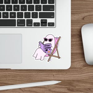 May include: A die-cut sticker featuring a light pink ghost wearing black sunglasses, sitting in a brown wooden beach chair while reading a purple book titled Spooky Summer with a palm tree and sunset graphic on the cover, accented by small white stars.
