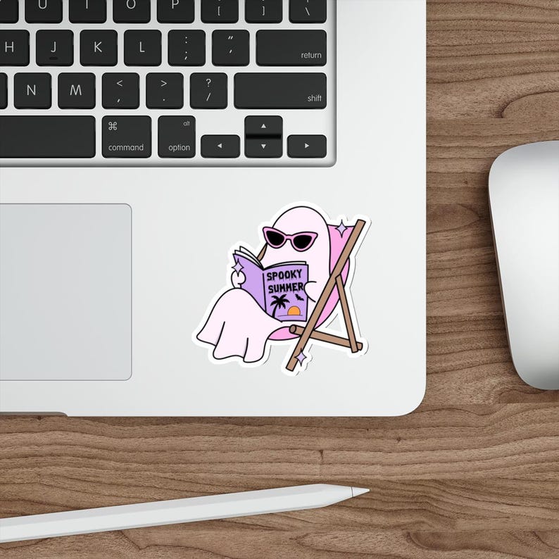 May include: A die-cut sticker featuring a light pink ghost wearing black sunglasses, sitting in a brown wooden beach chair while reading a purple book titled Spooky Summer with a palm tree and sunset graphic on the cover, accented by small white stars.