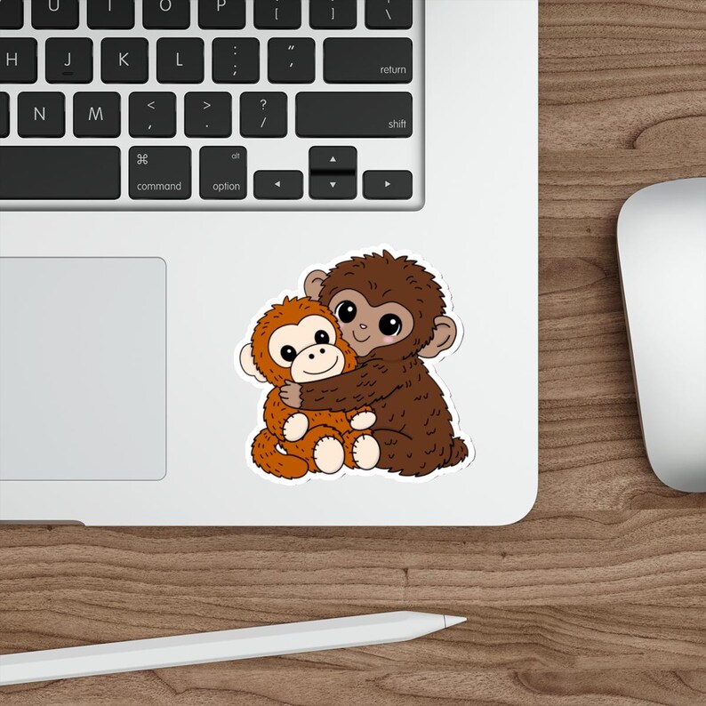 May include: A sticker featuring two cartoon monkeys hugging. One monkey is brown, and the other is orange. The sticker is on a white surface, with a laptop keyboard and a wooden desk in the background.