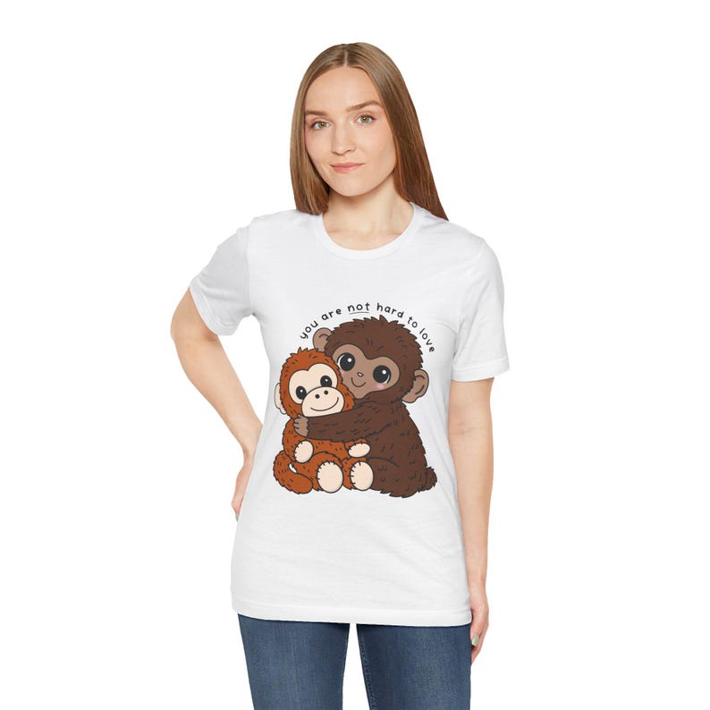 May include: White t-shirt featuring a cartoon illustration of two hugging monkeys. One monkey is brown, the other is orange. The text "you are not hard to love" is above the monkeys.