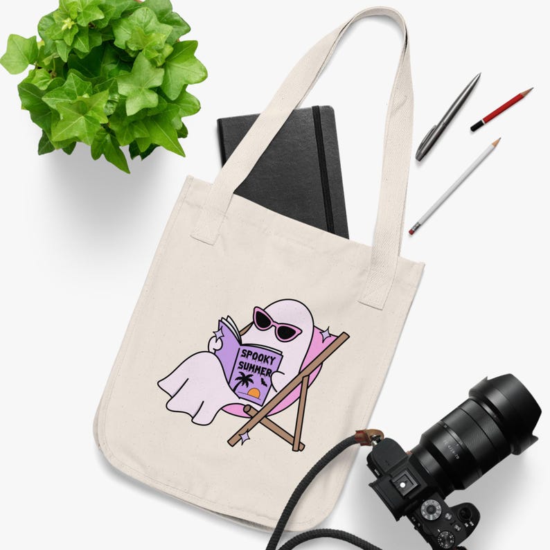 May include: A cream-colored canvas tote bag featuring a graphic of a light pink ghost wearing sunglasses, lounging in a beach chair while reading a book titled Spooky Summer with a palm tree and sunset illustration on the cover.