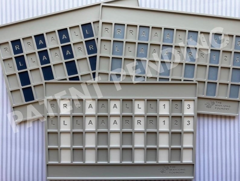 Op de afbeelding: Drie rechthoekige lichtgrijze plastic spelbordhouders met rastervakken. Elke houder heeft in reli&euml;f gedrukte letters R, A, L en cijfers 1, 2, 3 in de bovenste rijen. De onderste rijen bevatten lege vakken. Het Mahjong Foundry logo is rechtsonder zichtbaar.