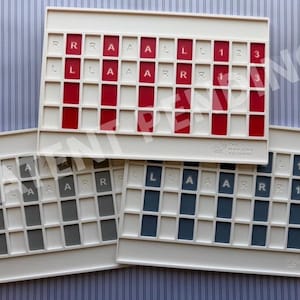 Op de afbeelding: Drie rechthoekige witte plastic spelrekken met rastervakken. Het bovenste rek heeft rode inzetstukken met witte letters R, L, A en cijfers 1, 2, 3. De onderste rekken hebben grijze en blauwe inzetstukken met identieke markeringen. Het Mahjong Foundry logo is zichtbaar.