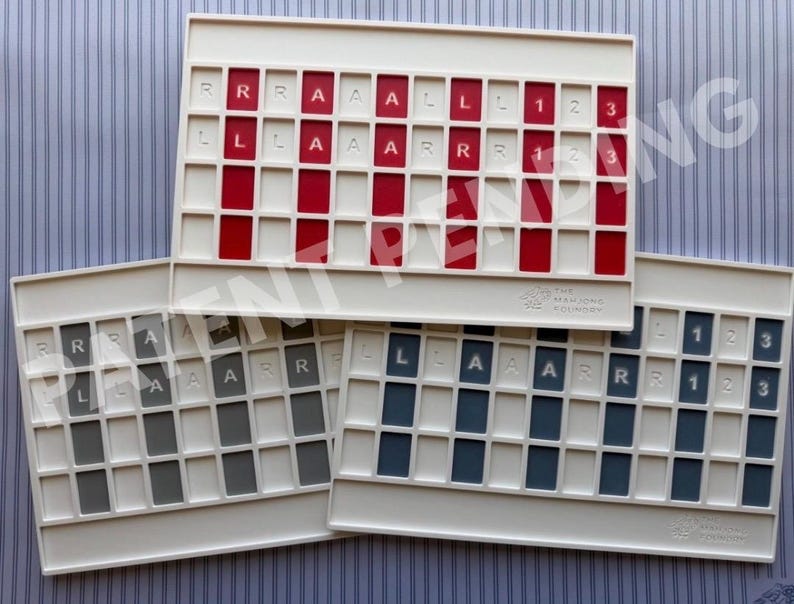 Op de afbeelding: Drie rechthoekige witte plastic spelrekken met rastervakken. Het bovenste rek heeft rode inzetstukken met witte letters R, L, A en cijfers 1, 2, 3. De onderste rekken hebben grijze en blauwe inzetstukken met identieke markeringen. Het Mahjong Foundry logo is zichtbaar.