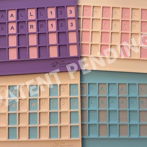 Op de afbeelding: Vier rechthoekige siliconen trays met rastervakken voor spelstenen. De trays zijn paars, cr&egrave;me, beige en blauw. Elke tray bevat in reli&euml;f aangebrachte letters A, L, R en cijfers 1, 2, 3 in specifieke vakken. Het Mahjong Foundry logo is zichtbaar.