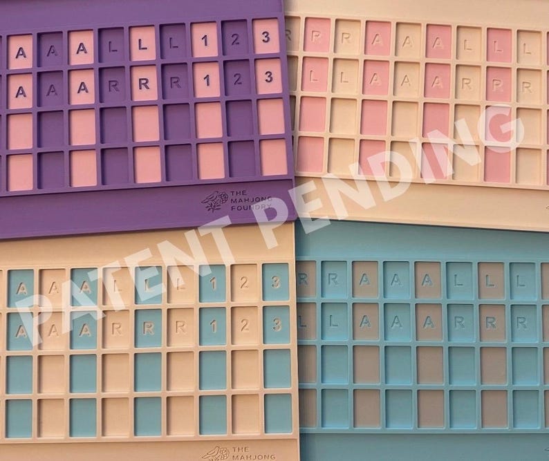 Op de afbeelding: Vier rechthoekige siliconen trays met rastervakken voor spelstenen. De trays zijn paars, cr&egrave;me, beige en blauw. Elke tray bevat in reli&euml;f aangebrachte letters A, L, R en cijfers 1, 2, 3 in specifieke vakken. Het Mahjong Foundry logo is zichtbaar.