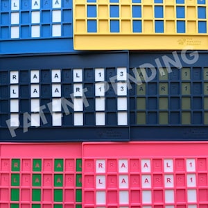Op de afbeelding: Een verzameling van zes rechthoekige mahjong-tegelhouders in blauw, geel, donkerblauw en roze. Elke houder heeft een raster van sleuven met witte, groene of donkere inzetstukken. De letters R, L, A en de cijfers 1, 2, 3 zijn op de inzetstukken gedrukt.