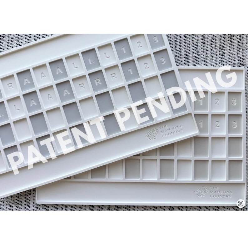 Op de afbeelding: Twee rechthoekige witte siliconen trays met een raster van verzonken vakjes voor het ordenen van tegels. De bovenste tray bevat in reli&euml;f aangebrachte letters en cijfers in specifieke cellen. Beide tonen het logo The Mahjong Foundry.