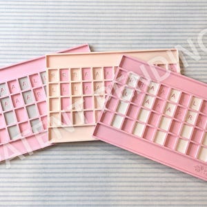Op de afbeelding: Drie rechthoekige siliconen trays in roze en perzik tinten, elk voorzien van een raster met vierkante vakjes. Sommige vakjes bevatten reli&euml;fletters en cijfers. Op de onderste rechter tray staat de tekst The Mahjong Foundry.