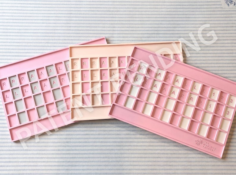 Op de afbeelding: Drie rechthoekige siliconen trays in roze en perzik tinten, elk voorzien van een raster met vierkante vakjes. Sommige vakjes bevatten reli&euml;fletters en cijfers. Op de onderste rechter tray staat de tekst The Mahjong Foundry.