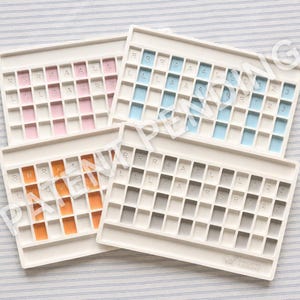 Op de afbeelding: Vier rechthoekige witte kunststof trays met rastervakken voor het organiseren van speelstenen. Elke tray heeft gekleurde inzetstukken in roze, blauw, oranje of grijs. Sommige vakken bevatten de letters R, A, L of de cijfers 1, 2, 3. Het Mahjong Foundry logo is zichtbaar.