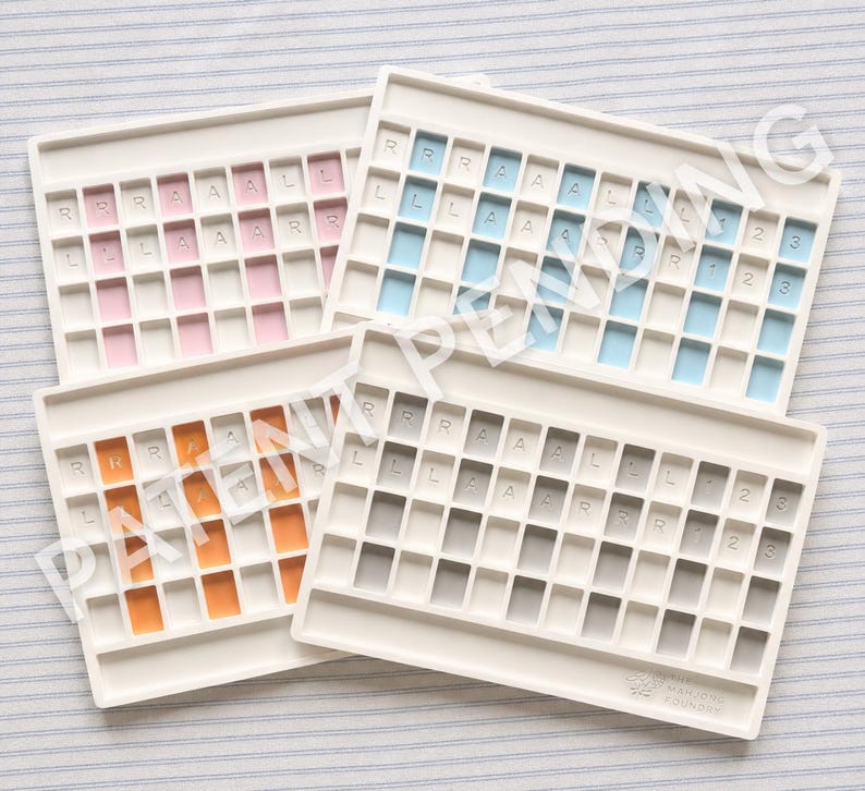 Op de afbeelding: Vier rechthoekige witte kunststof trays met rastervakken voor het organiseren van speelstenen. Elke tray heeft gekleurde inzetstukken in roze, blauw, oranje of grijs. Sommige vakken bevatten de letters R, A, L of de cijfers 1, 2, 3. Het Mahjong Foundry logo is zichtbaar.